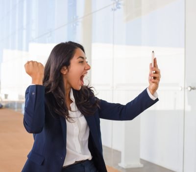 Excited young woman holding smartphone. Emotional businesswoman using mobile phone and shaking fist. Connection concept