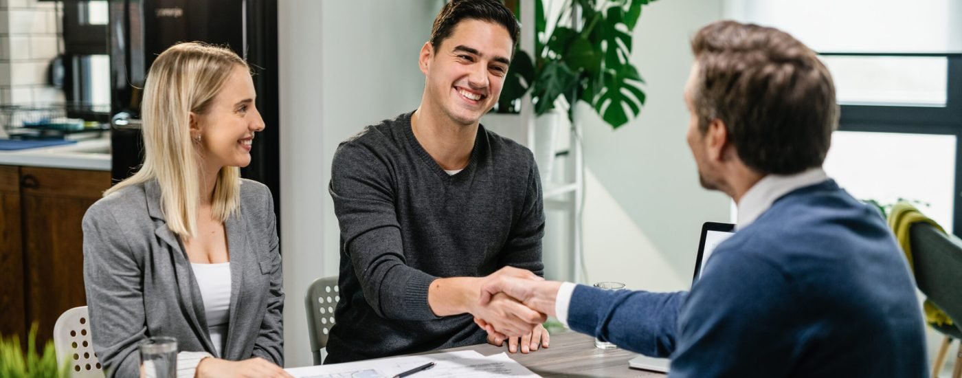 Young happy couple making an agreement with their financial advisor at home. Men are shaking hands.